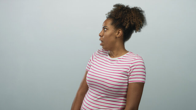 Young african american woman mouth open looking sideways in studio with light blue backdrop wearing pink striped tee and hair in curly puff; surprise reaction.