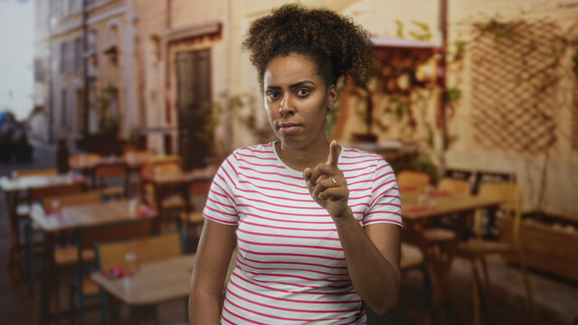 Woman points finger at camera in outdoor restaurant terrace, wearing striped shirt and ring on finger, curly hair up, standing near tables and chairs; angry warning.