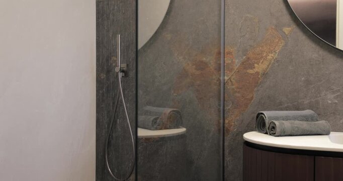 Walk-in shower and premium stone bathroom interior, static video captures the first water streams descending beside a rounded wood vanity and large round mirror in an elegant bathroom setting