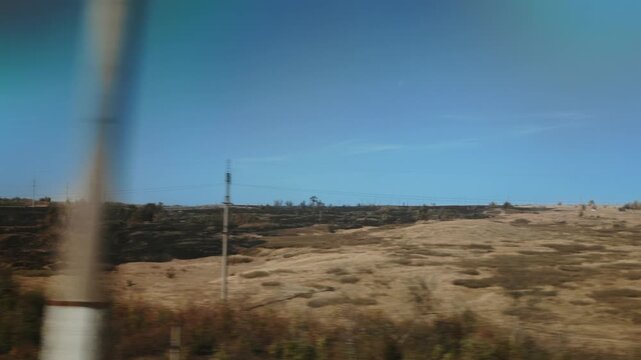 Panoramatic view of extensive rural terrain from a speeding vehicle, showcasing areas of charred earth adjacent to dry golden fields, utility infrastructure lining horizon, bright blue sky above