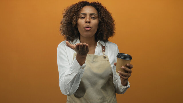 Young african american woman baker in apron holding a takeaway coffee cup and blowing kiss in studio; hospitality joy.