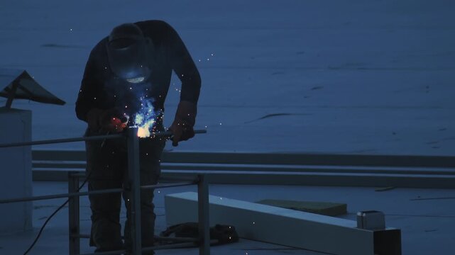 Professional welder in protective gear carefully joins metal components on flat roof surface at dusk, orange sparks illuminate work area highlighting industrial activity. Industrial process, hard work