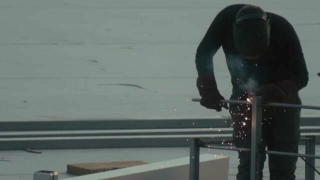 Industrial worker engaged in arc welding process on roof structure. Person wears full protective gear including welding mask and heavy gloves. Bright sparks fly from point of contact, smoke and light 