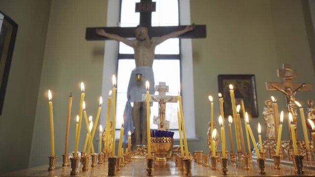 Close up view of burning yellow wax candles in rows on golden holders on altar table, casting soft light. Out of focus wooden crucifix of Jesus Christ visible in distance.