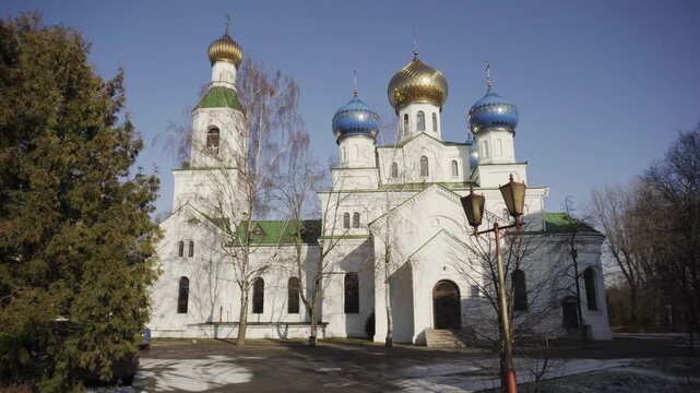 Exterior view of majestic white Orthodox church. Five prominent domes: large golden dome, two smaller golden domes, two blue domes with golden stars. Bell tower with golden cupola. Green roofs, bare t