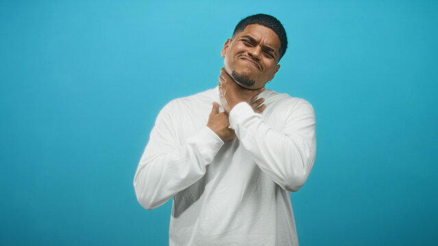 Hispanic man clutching throat with both hands in a blue studio backdrop, grimacing and gagging; pain struggle.