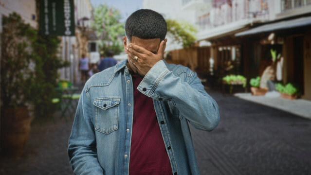 Hispanic man with beard holding eyeglasses and rubbing eyes in a cobbled street wearing denim jacket and maroon shirt; fatigue inconvenience.