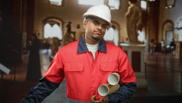 Man construction worker in white hard hat and red uniform holding rolled blueprints and tilting his head in a museum gallery; restoration duty.
