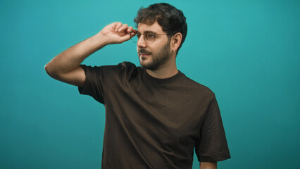 Bearded man adjusts glasses with right hand in studio against teal backdrop wearing brown t shirt and hoop earring  contemplation calm introspection. © Krakenimages.com