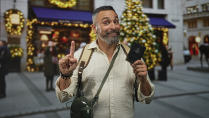 Naklejka premium Man pointing finger while holding passport on a decorated street with christmas tree and lights; holiday travel joy.