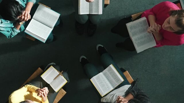 Top view of skilled diverse prayer clasping hand and praying to god while sitting at chair in circle. Top down aerial view of people folded hand while sitting with opened bible on laps. Symposium.