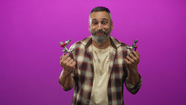 Man holding two faucet valves with both hands in a magenta studio, wearing a flannel shirt with grey beard and short grey hair; playful repair.