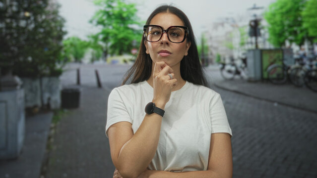Woman with long brunette hair wearing white t shirt and oversized glasses, smartwatch visible, hand on chin on a cobblestone street with bicycles nearby; contemplation.