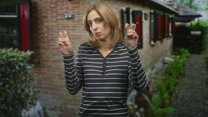 Naklejka premium Woman blonde showing air quotes with both hands and pursed lips in front of a brick building on a garden path, gesture ironic; wry sarcasm.
