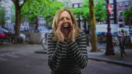 Fototapeta premium Woman cupping hands and shouting with open mouth on an urban street near bicycles and trees, wearing striped long sleeve top; alarm protest anger.