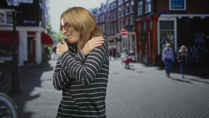 Fototapeta premium Woman hugs herself with crossed arms wearing glasses and striped sweater on a sunlit street; self comfort serenity.