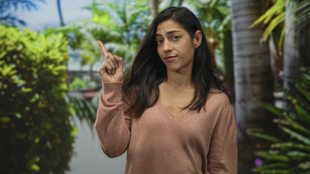 Young hispanic woman points index finger upward in a green park with palm trees and dense foliage while wearing a v neck sweater; confidence persuasion.