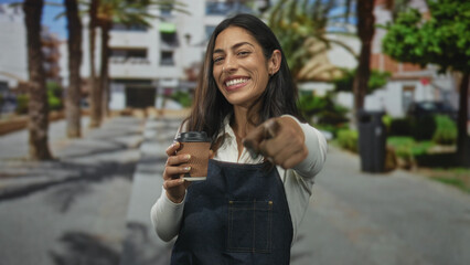 Fototapeta premium Woman points finger at viewer while holding takeaway coffee cup wearing denim apron on street; welcoming local cafe.