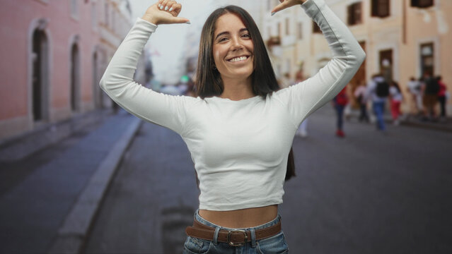 Young woman showing midriff in a white long sleeve top, thumbs pointing to chest, smiling in a busy street with pedestrians; confidence joy celebration.