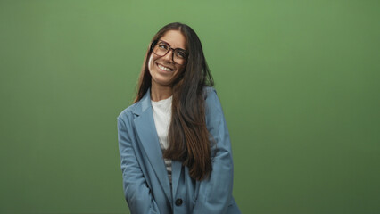 Obraz premium Woman with glasses smiling, hand to mouth and arms crossed wearing blue blazer in a green studio; confidence charm.