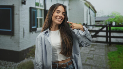 Fototapeta premium Young hispanic woman points fingers to cheeks and smiles in front of building by a wooden gate and stone path; joy confidence.
