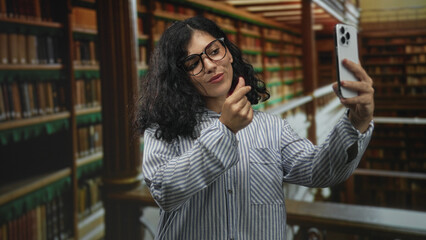 Fototapeta premium Young woman holding smartphone and making finger heart gesture in building library; self expression confident.