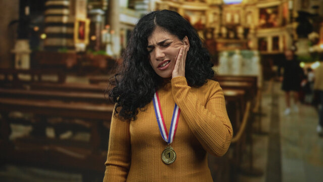 Young woman holding cheek and wincing with gold medal on chest, wearing mustard sweater inside a church building with pews and blurred worshipers; toothache pain.
