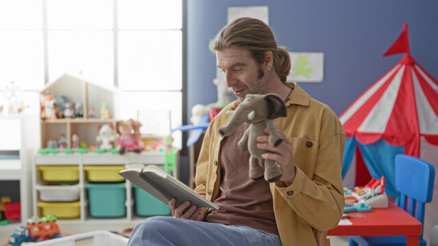 Man holds a stuffed elephant puppet and reads aloud from an open book while seated in a classroom play area with toys and storage bins; nurturing storytelling connection.