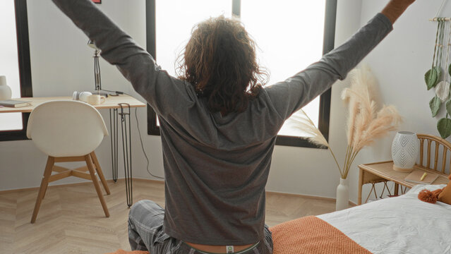 Man on bed sits at edge and stretches both arms upward toward window with desk and pampas grass in bedroom; morning calm.