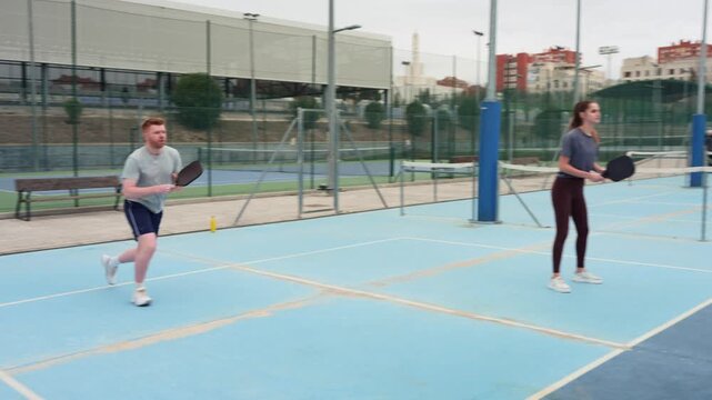 Two young friends, a man and a woman, enjoying a game of pickleball. The man serves the ball while his partner awaits the return