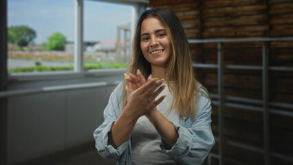 Fototapeta premium Woman clapping hands together in a well lit indoor studio near a large window and wooden ladder wall, smiling toward camera; joy.