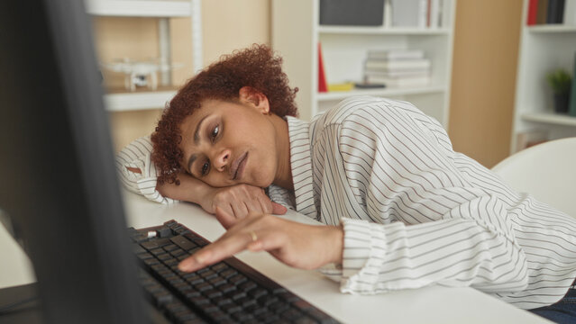 Woman resting head on desk and reaching for keyboard beside monitor in office building, leaning forward with slumped posture and blank expression; fatigue.