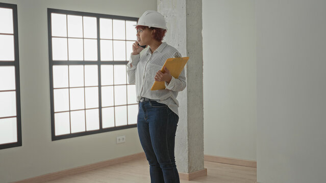 Woman architect wearing hardhat holds phone to ear while holding envelope folder in building windowed room; project planning focused.
