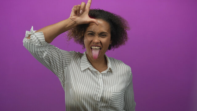Woman sticking out tongue and making loser l sign with hand on forehead in a purple studio; playful mischief.