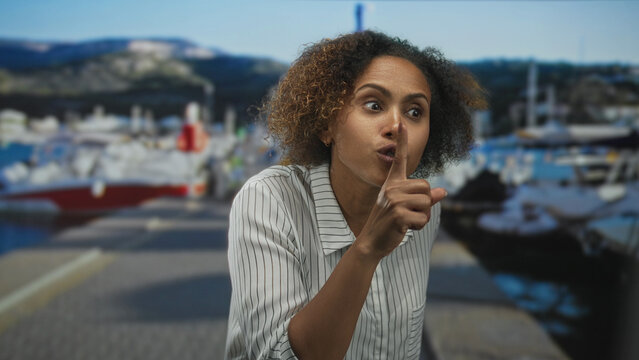 Woman with finger to lips for silence at a seaside port pier, leaning forward in a shushing gesture; silence secrecy serious.