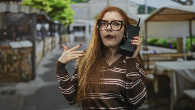 Redhead woman in striped sweater holds a large vintage brick phone to ear and gestures with her hand on a street market; frustration.