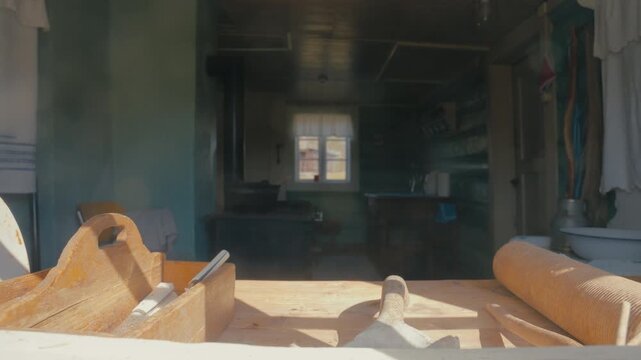 Warm sunlight falls across old baking tools on a wooden table, while a dim rustic kitchen recedes behind them, creating a nostalgic domestic scene.