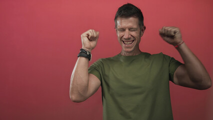 Naklejka premium Man smiling and raising both fists, showing forearms and bracelets against a vivid red studio wall; joy celebration.
