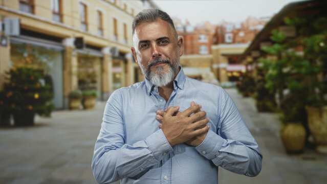 Middle aged man with gray beard holding hands to chest on a street lined with shops and potted trees, smiling gently; gratitude.