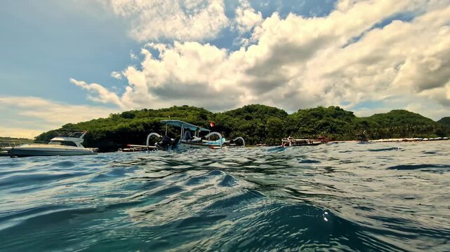 A water surface perspective after scuba diving in Bali, showing a traditional jukung boat with Indonesian flag, a speedboat and a lush green tropical island coastline under a dramatic cloudy sky.