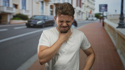 Fototapeta premium Young hispanic man clutching his neck and grimacing while standing on a city street wearing a white t shirt and squinting in daylight; sore throat pain.