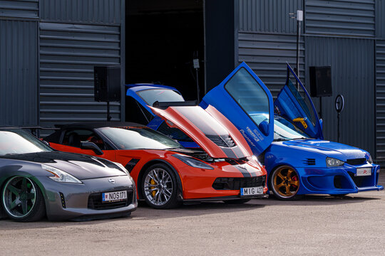 Group of modified performance cars including a grey Nissan 370Z, orange Chevrolet Corvette, and blue Volkswagen Golf with vertical scissor doors at a custom automotive show