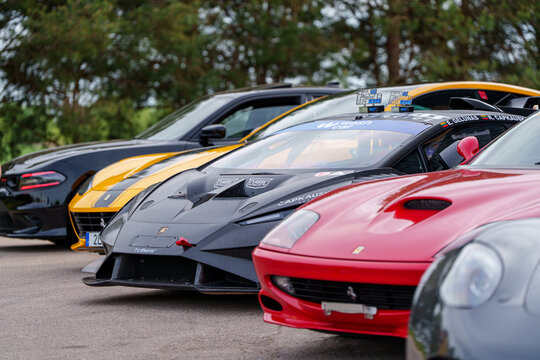 Line of luxury supercars including a matte black Lamborghini, red Ferrari, and yellow Ferrari 812 parked at a high-end automotive race event or car meet