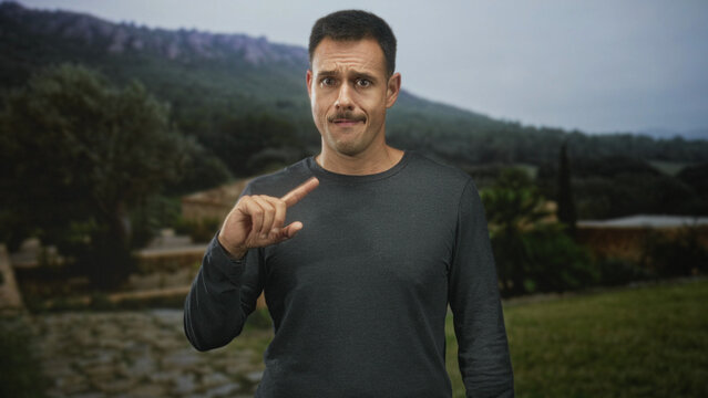 Man with moustache pointing index finger beside a stone path near a villa in forest, uncertain expression; hesitation.