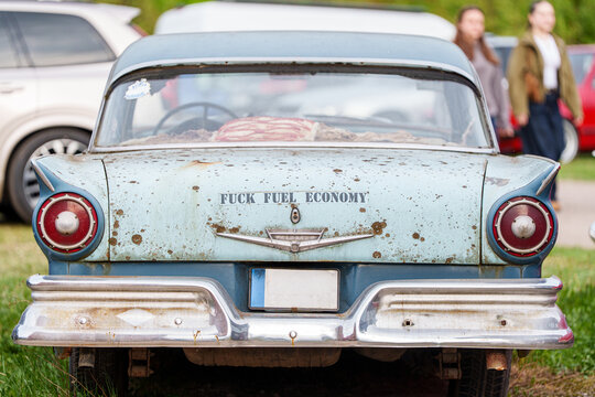 Rear view of a vintage 1957 Ford Fairlane with heavy patina and rust, featuring a provocative fuel economy decal on the trunk, classic American oldtimer in weathered condition