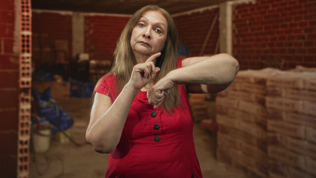 Woman pointing finger to wrist in unfinished brick building with stacked pallets and tools; impatience.