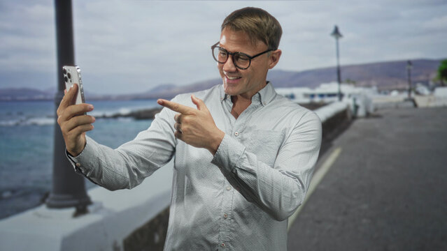 Man holding smartphone for a selfie with hand on chest on a seaside street wearing glasses and button shirt; sincere connection.