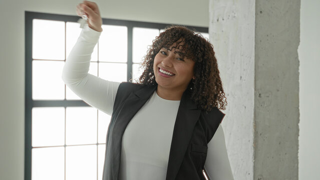 Woman with curly brunette hair holds keys high with hand raised, smiling in empty building interior by window and concrete pillar; new home joy.