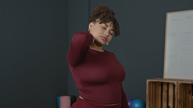 Woman massaging neck and stretching shoulder in studio with yoga mat and exercise balls; relief recovery wellness.