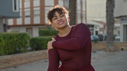 Woman rubbing shoulder with eyes closed and a grimace on a sunlit street near buildings, wearing a burgundy top and hoop earrings  relief recovery. © Krakenimages.com